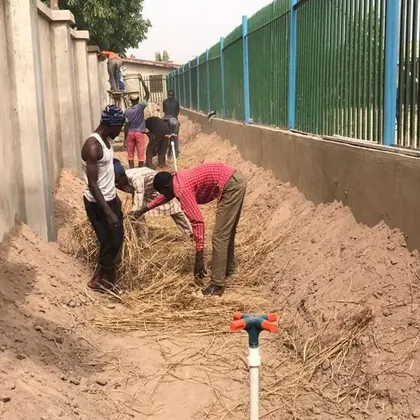 People digging a long planting trench beside a wall, with an irrigation pipe in place.