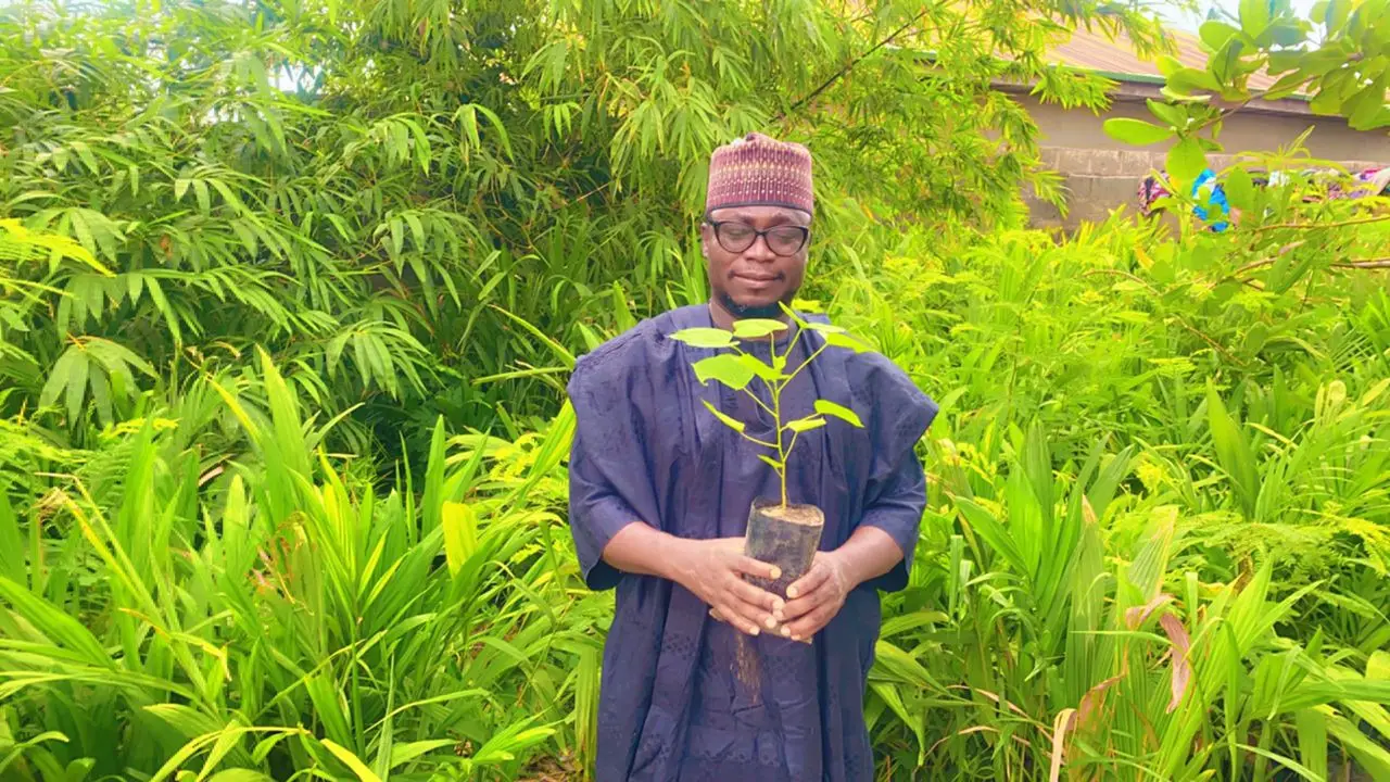A man in a navy kaftan and embroidered cap holds a tree seedling in a nursery bag, standing in dense green foliage.